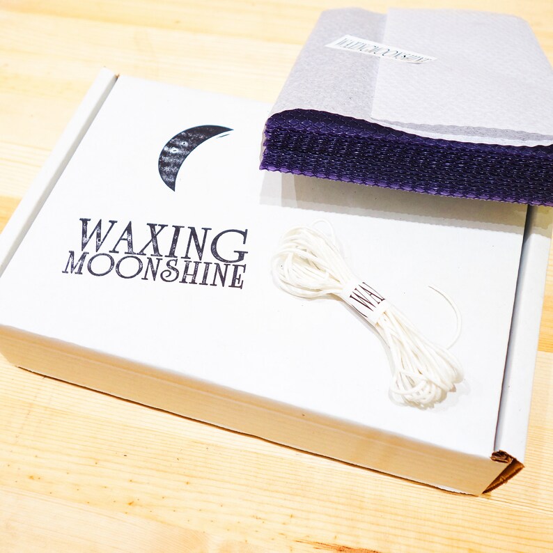 May include: A white box with the text "WAXING MOONSHINE" and a crescent moon logo. The box contains purple sheets, and a bundle of white string. The items are on a light wood surface.