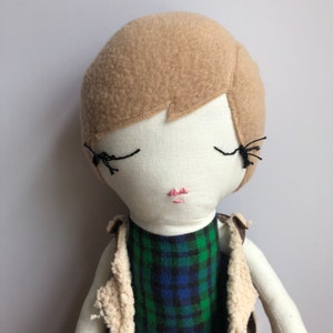 Puede incluir: Una muñeca hecha a mano con cabello castaño, ojos cerrados y una boca cosida de color rosa. La muñeca lleva una camisa a cuadros verde y azul y un chaleco de piel sintética marrón.