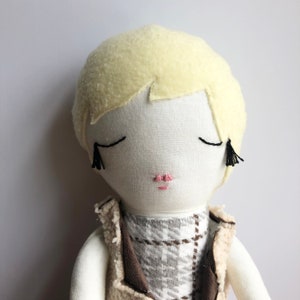 May include: A handmade doll with blonde hair, closed eyes, and a pink embroidered mouth. The doll is wearing a brown and white plaid shirt and a brown vest.