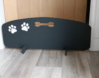 May include: A black wooden pet gate with white paw prints and a bone cutout. The gate has two support legs and is designed to stand on a wooden floor. The gate is a decorative way to keep pets contained.