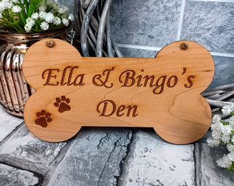 May include: A wooden dog bone-shaped sign with the text "Ella & Bingo's Den" engraved on it. The sign has two paw prints carved into the wood.