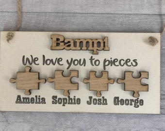 May include: Wooden sign with the word "Bampi" at the top, and the phrase "We love you to pieces" below. Four wooden puzzle pieces are below the phrase, with names Amelia, Sophie, Josh, and George beneath them. A jute rope is attached for hanging.
