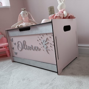 May include: A pink wooden toy chest with a silver trim and the name "Olivia" written on the front. The chest has a fairy and butterfly decorations.