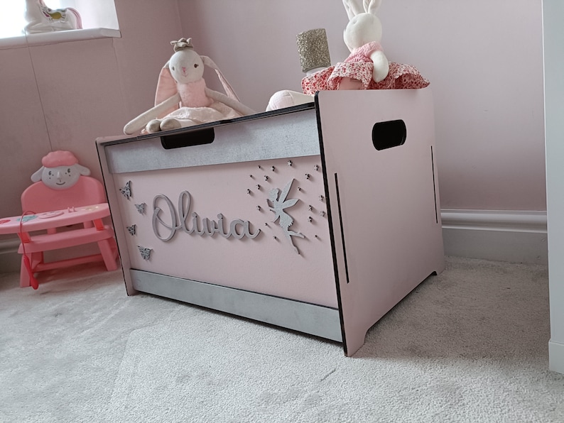 May include: A pink wooden toy chest with a silver trim and the name "Olivia" written on the front. The chest has a fairy and butterfly decorations.