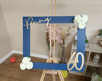 May include: A navy blue photo booth frame with the name "Penny" in gold script and the number "60". White roses adorn two corners. The frame is displayed on a wooden easel, with floral arrangements in the background.