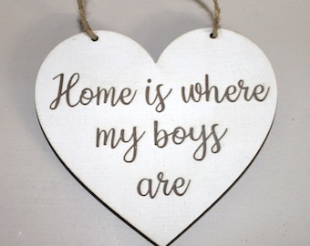 May include: A white wooden heart-shaped sign with the phrase "Home is where my boys are" in a cursive font. The sign has a natural twine loop for hanging.
