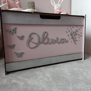 May include: A pink wooden toy chest with a gray trim and a silver glitter finish. The chest has a cutout of a fairy with a dandelion, the name "Olivia" and several butterflies.