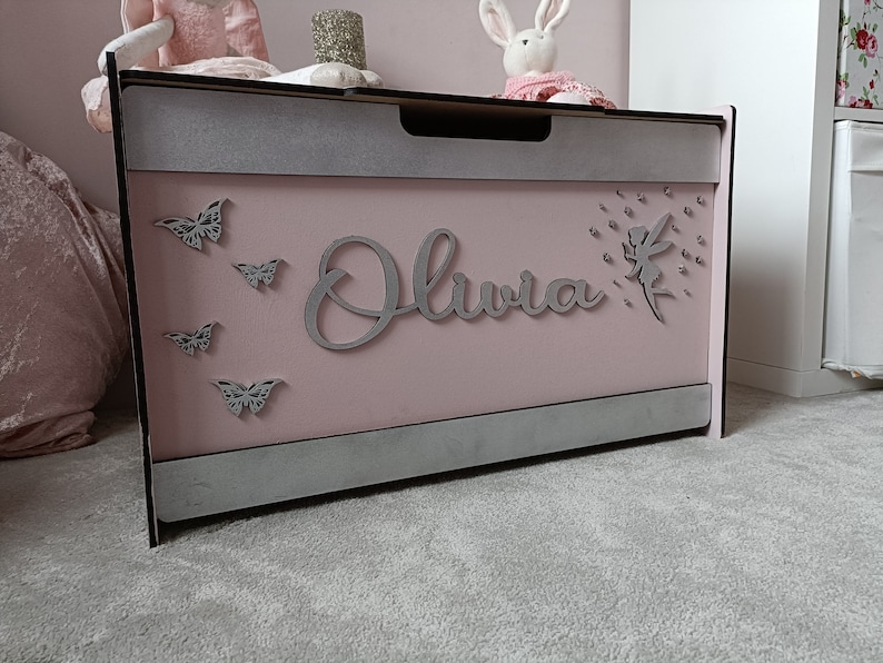 May include: A pink wooden toy chest with a gray trim and a silver glitter finish. The chest has a cutout of a fairy with a dandelion, the name "Olivia" and several butterflies.