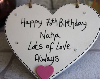 Może przedstawiać: Białe drewniane serce z czarnym napisem "Happy 70th Birthday Nana Lots of Love Always". Serce ma przyklejone małe różowe serce.