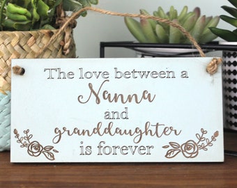 May include: A light blue wooden sign with the text "The love between a Nanna and granddaughter is forever" in brown lettering. The sign has a rustic twine hanger and is decorated with two small floral designs.