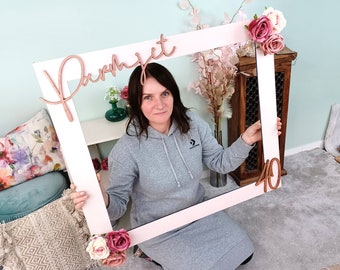 May include: A white photo booth frame with a pink background. The frame has a wooden sign that says "Parmjeet" and a wooden number "40". The frame is decorated with pink and white flowers.