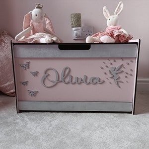 May include: A pink toy chest with a silver glitter trim and the name "Olivia" written on the front. The chest is decorated with silver butterflies and a fairy. Two plush toys, a bunny wearing a crown and a bunny wearing a dress, are sitting on top of the chest.
