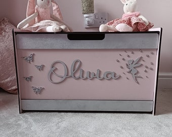 May include: A pink toy chest with a silver glitter trim and the name "Olivia" written on the front. The chest is decorated with silver butterflies and a fairy. Two plush toys, a bunny wearing a crown and a bunny wearing a dress, are sitting on top of the chest.