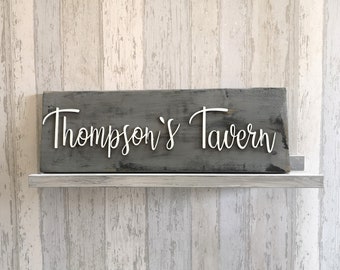 Może przedstawiać: Rusztykalny drewniany znak z napisem "Thompson's Tavern" w białych wypukłych literach. Znak ma postarzaną szarą powierzchnię i idealnie nadaje się do baru lub restauracji.