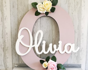 May include: A pink wooden letter O with the name Olivia written in white cursive letters. The letter is decorated with white and pink artificial flowers.