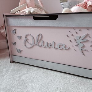 May include: A pink wooden toy chest with a silver trim and the name "Olivia" written on the front. The chest is decorated with silver butterflies and a fairy with stars.