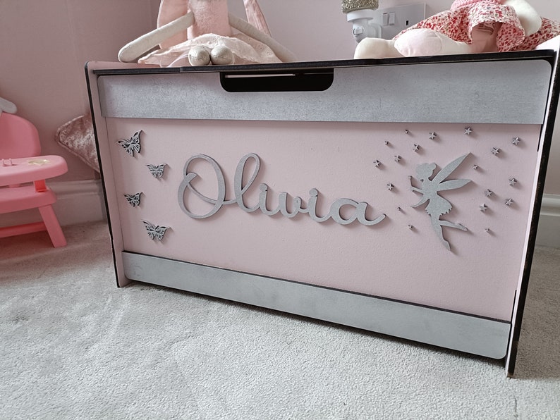May include: A pink wooden toy chest with a silver trim and the name "Olivia" written on the front. The chest is decorated with silver butterflies and a fairy with stars.