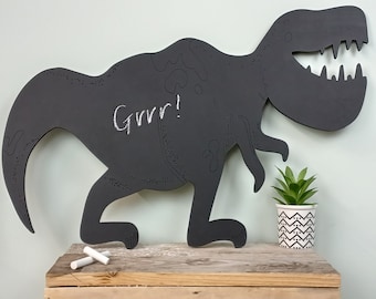 May include: A black chalkboard shaped like a dinosaur, with the word "Grrr!" written in white chalk. The dinosaur is standing on a wooden surface with two pieces of white chalk next to it.