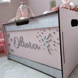 May include: A pink wooden toy chest with a silver trim and a silver fairy cutout on the front. The chest has the name "Olivia" written on the front in silver letters. There are also silver butterflies and stars on the front of the chest.