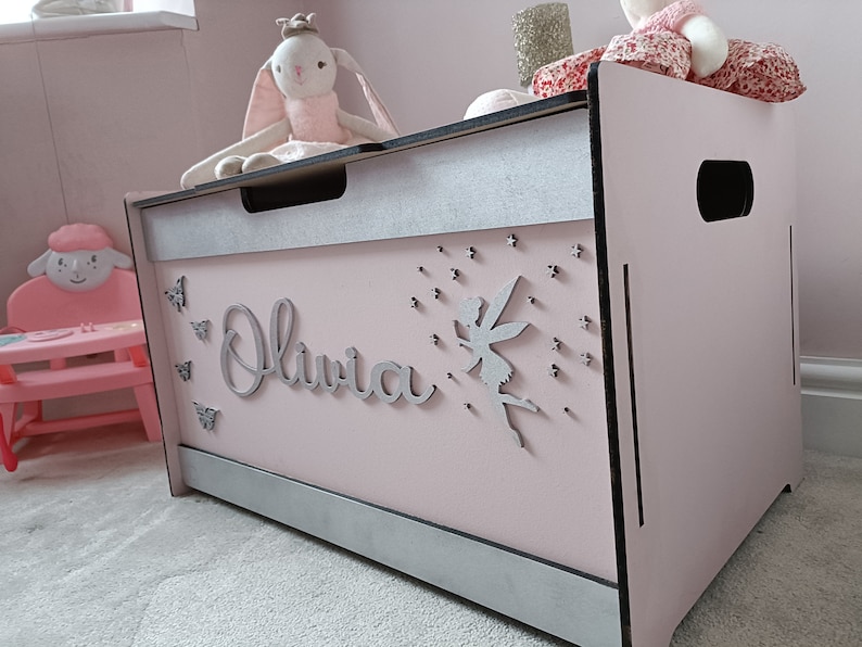 May include: A pink wooden toy chest with a silver trim and a silver fairy cutout on the front. The chest has the name "Olivia" written on the front in silver letters. There are also silver butterflies and stars on the front of the chest.