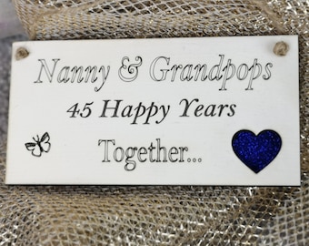 Może przedstawiać: Biały drewniany znak z tekstem "Nanny & Grandpops 45 Happy Years Together..." z niebieskim sercem z brokatem i grafiką motyla.
