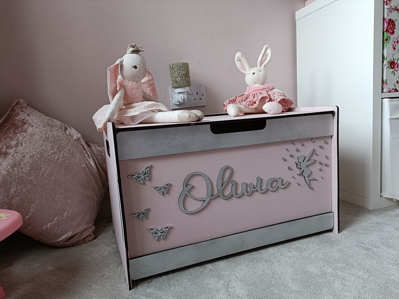May include: A pink toy chest with a silver trim and the name "Olivia" written on the front. The chest is decorated with silver butterflies and a silver fairy with a sparkly dress. Two plush bunny toys are sitting on top of the chest.