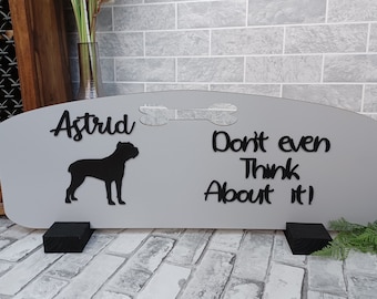 May include: A gray sign with the name "Astrid", a dog silhouette, and the phrase "Don't even Think About it!" in black lettering. The sign has a bone-shaped cutout and is supported by two black blocks.