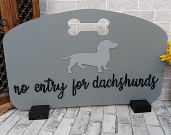 Może przedstawiać: Szary drewniany znak z wycięciem w kształcie kości i sylwetką psa rasy dachshund. Na znaku widnieje napis "no entry for dachshunds".