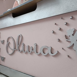 May include: A pink wooden toy chest with a silver lid and trim. The chest has a silver fairy silhouette with wings and stars, and the name "Olivia" is written in silver letters.