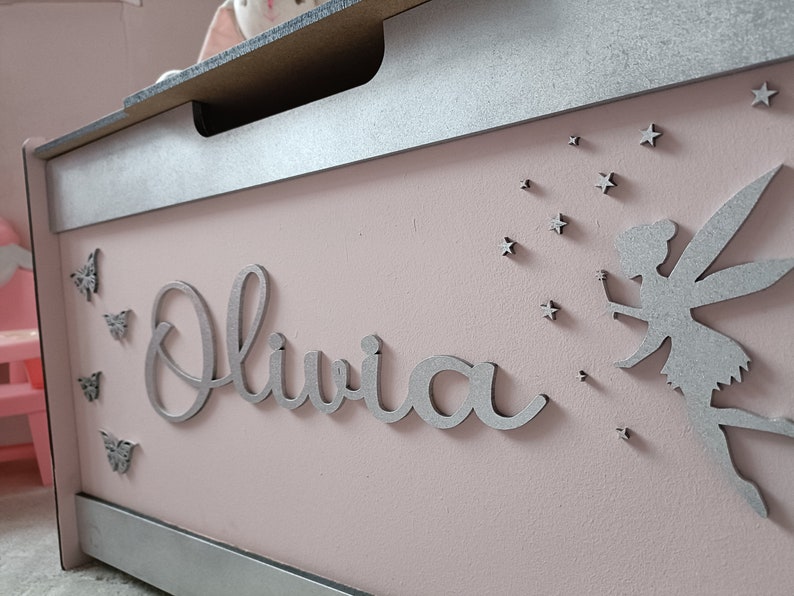 May include: A pink wooden toy chest with a silver lid and trim. The chest has a silver fairy silhouette with wings and stars, and the name "Olivia" is written in silver letters.
