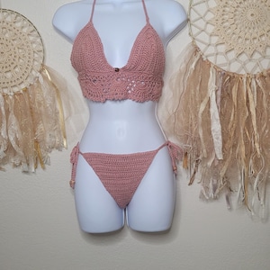 May include: A handmade, dusty rose crochet bikini set displayed on a white mannequin. The top is a halter style with a decorative trim, and the bottoms are a classic triangle cut. The set is displayed with two dream catchers in the background.