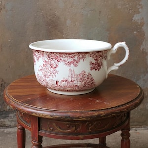 May include: A white ceramic bowl featuring a red toile design of a landscape. The bowl has a handle and sits on a small, round, wooden table. The table is painted red. The bowl's diameter is approximately 25 cm.