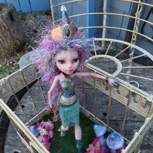 May include: A fantasy-themed custom doll. The doll features a detailed face, purple and blue hair, and a teal outfit with gold-coloured belt and accessories. The doll is positioned inside a decorative metal basket.
