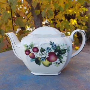 Puede incluir: Tetera de porcelana blanca con tapa y ribete dorados. La tetera presenta un diseño de frutas con manzanas, bayas y peras. El asa y el pico son curvos. Fondo borroso con follaje otoñal.