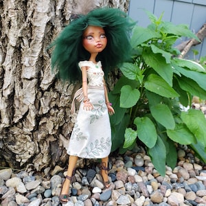 May include: A doll with voluminous dark green hair, wearing a white off-the-shoulder top with a crochet-style bodice and a matching skirt featuring a green leaf pattern. The doll has brown skin, brown eyes, and wears brown strappy sandals.