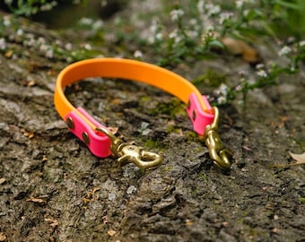 May include: An orange and pink dog leash with brass-colored hardware. The leash is made of a flexible material and is resting on a textured, brown surface. The leash has a bright, eye-catching color scheme.