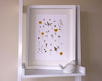 May include: A white framed print with a white background and pressed flowers in shades of yellow and white. The print is on a white shelf with a white ceramic bird figurine.