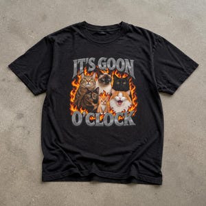 Könnte beinhalten: Ein schwarzes Kurzarm-T-Shirt mit einem Grafikdruck von sechs Katzen, die von orangefarbenen und gelben Flammen umgeben sind. Der Text IT'S GOON O'CLOCK ist in einer metallisch silbernen Schrift über und unter den Katzen mittig aufgedruckt.