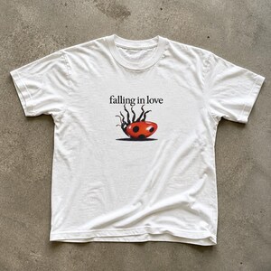 May include: A white short-sleeved t-shirt featuring a graphic of a red ladybird with black spots lying on its back with its legs in the air. Above the ladybird, the text "falling in love" is printed in a black, serif-style font.