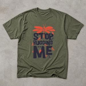 Può includere: Una t-shirt a maniche corte color verde oliva con una stampa grafica di una grande libellula arancione sopra il testo blu scuro "STOP BUGGING ME" e piccole illustrazioni di libellule arancioni sparse attorno alla scritta sul davanti.