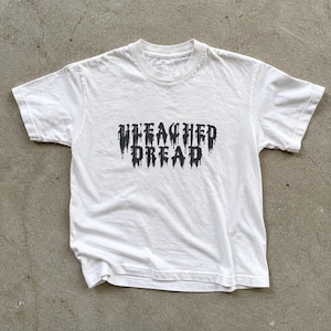 Peut inclure: Un t-shirt blanc à manches courtes avec le texte "UNLEASHED DREAD" imprimé en noir dans une police gothique dégoulinante au centre de la poitrine. Le vêtement présente un col rond standard et un dos ainsi que des manches unis et blancs.