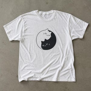 Könnte beinhalten: Ein weißes T-Shirt mit kurzen Ärmeln und einem zentralen Grafikdruck eines Yin-Yang-Symbols, das aus zwei stilisierten Katzen besteht. Eine Katze ist weiß mit schwarzen Umrissen, die andere schwarz mit weißen Umrissen, beide mit geschlossenen Augen.