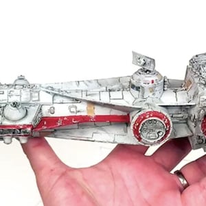 Könnte beinhalten: Detailliertes Modell eines Raumschiffs, überwiegend weiß mit roten und grauen Akzenten. Das Modell weist komplizierte Verkleidungen, Türme und Motordetails auf. In einer Hand gehalten, ist das Modell etwa 30 cm lang und zeigt eine verwitterte Ästhetik.