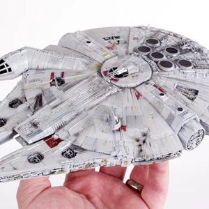 Op de afbeelding: Een gedetailleerd model van het Millennium Falcon ruimteschip, in lichtgrijs met rode en zwarte accenten. Het model heeft ingewikkelde details, waaronder panelen, ventilatieopeningen en een cockpit. Het iconische ruimteschip is een verzamelobject.