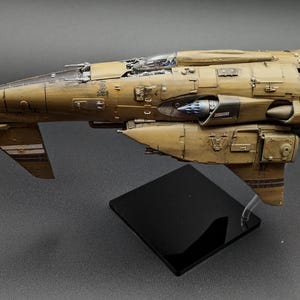 May include: A detailed model spacecraft, predominantly tan and brown, with intricate paneling and design. It features wings, thrusters, and a cockpit, mounted on a black base. The design suggests a science fiction or fantasy theme, with a focus on detail.