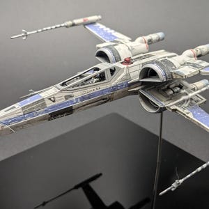 May include: A detailed model of a Star Wars X-wing starfighter. The spacecraft is primarily white with blue accents on the wings and fuselage. The model is mounted on a stand, showcasing its design and features.