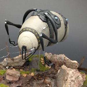 May include: A detailed model of a robotic device with a cylindrical body in off-white and black. The model has articulated legs and a series of black, curved appendages. Accents include blue and yellow details. The model is set on a diorama with rocks and greenery.