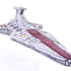 Puede incluir: Modelo detallado de una nave espacial, predominantemente blanco con detalles rojos y grises. La nave presenta un cuerpo largo y rectangular con estructuras en forma de ala y una torre prominente. El modelo se exhibe sobre un soporte transparente.