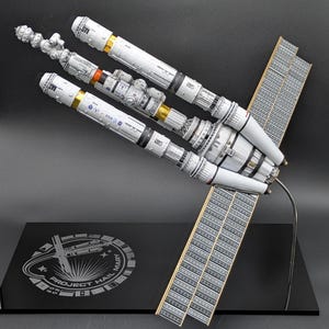 May include: A detailed scale model of the Astrophage-powered spaceship from Project Hail Mary, featuring white cylindrical modules, gold foil accents, and large rectangular solar arrays. The model is mounted on a black base with the text Project Hail Mary engraved in a circular emblem.