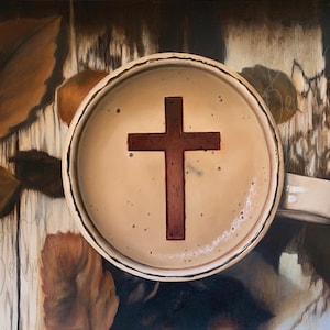 May include: A coffee cup with a cross design in the coffee. The cup is white with a handle, and the coffee is light brown. The cross is a darker shade of brown. The background includes brown leaves and a wooden surface.